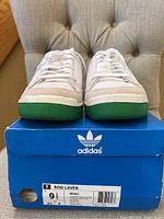 Front view of sneakers on original blue Adidas box showing green rubber toe caps and white mesh upper.
