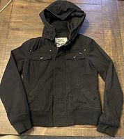Full view of the black Aritzia TNA hooded jacket laid flat, showing front design details including pockets, snap buttons, and ribbed edges.