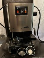 Front view of espresso machine on black cloth with portafilter, two filter baskets and scoop in foreground