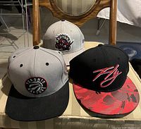 Three baseball caps placed on a striped chair: two Toronto Raptors caps (one grey with black brim, one grey with embroidered dinosaur logo) and a black cap with 'Fly' embroidery and red patterned brim.