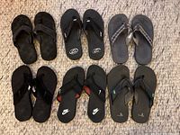 Lot of six pairs of men's black flip flops displayed on woven mat, viewed from top, showing different strap styles and wear