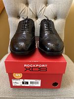 Front view of the black leather dress shoes on top of the red Rockport XCS box, showing wingtip brogue details and lace-up design.