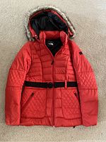 Full view of the red North Face women's coat, showing front side with belt and fur-trimmed hood on carpet background.