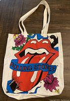 Front side of tote bag with colorful Rolling Stones tongue logo, blue ribbon with text, and floral accents.