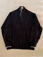 Full view of black sweater laid flat showing front, long sleeves, ribbed cuffs and hem, with Tommy Hilfiger logo on chest.