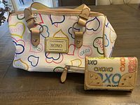 Xoxo purse and wallet set displayed on wooden table, showing colorful heart and XOXO text print, purse front with tan leather handles and logo plaque, wallet front with logo and stud details.