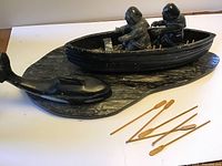 Close view of soapstone boat with two carved figures on whale-shaped base, six wooden oars set nearby