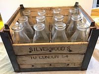 Photo showing wooden milk crate with 12 glass milk bottles, marked Silverwood's 10 London 54 on crate side.