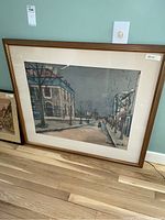 Framed art print showing an outdoor street scene with buildings, bare trees, and a figure walking. Signed lower right. Wood frame and cream matting visible.