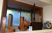 Shelf with a set of wooden bookends including a hand-carved rhino head and a plain wooden block. Also includes a wooden turned post and a kitchen scale with dial.