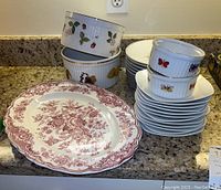 Photo showing a pile of porcelain appetizer plates decorated with butterflies and floral motifs next to berry-themed baking dishes and a large red floral patterned platter on countertop.