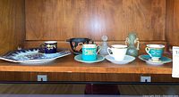 Shelf holding an assortment of vintage demitasse and espresso cups, one with black and gold pattern, a couple with teal and gold designs, a decorated plate, a green bud vase, and a crystal perfume bottle. Also visible is a redware owl-shaped bowl.