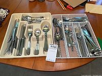 Overview of flatware items arranged in two trays on wooden table, showing knives, forks, spoons, and serving utensils.