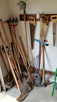 Photo showing assortment of long-handled gardening and yard tools including spades, sledgehammer, aerator, cultivators, rakes, and hand saws hung on wall and leaning in corner.
