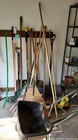 Photo showing assorted gardening tools including rakes, shovels, and brooms against a wall and floor.