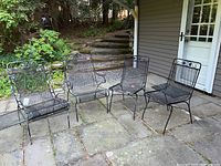 Wide view of all four wrought iron patio chairs arranged on stone patio. One chair has visible pull-out tray under seat.