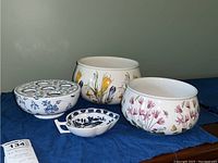 All four ceramic pieces displayed on a dark blue fabric: two large Portmeirion bowls (one with crocus yellow/purple flowers, one with pink cyclamen flowers), a smaller blue and white Mottahedeh bowl, and small nut dish with blue floral patterns and handle.