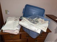 Stack of folded table linens and napkins, including lace and fabric pieces in light colors, on a wooden dresser next to plastic containers.