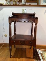 Front view of vintage wooden washstand showing drawer with two wooden knobs, turned legs, scalloped apron, and raised back and sides on the top surface.