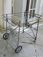 Three-quarter front view of metal bar cart showing top glass shelf, circular frame details and large rear wheels