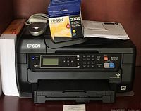 Full view of black Epson WorkForce WF-2750 printer with control panel and LCD screen lit. Ink cartridge package and manual placed on top of printer.