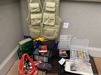 Front view of beige mesh fishing vest hanging with blue-yellow duffel and assorted tackle boxes and bags beneath