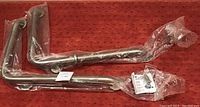 Two L-shaped stainless steel safety grab bars wrapped in protective plastic on a carpeted surface, showing length proportions and mounting brackets.