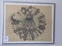 Framed artwork showing circular pattern of stylized face masks in black ink on beige paper, signed by artist.