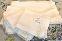 Stack of white hemstitched linen-cotton blend napkins and placemats on floral tablecloth with auction label on top.
