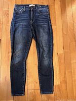 Full view of blue skinny jeans laid flat on wood floor, showing front side with button and zipper closure, five pockets, and slight fading on legs.