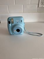 Front view of light blue Fujifilm Instax Mini 11 instant camera showing lens, flash, and viewfinder.