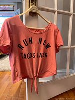 Coral-pink short sleeve active top with front tie knot and black text 'RUN NOW TACOS LATER'