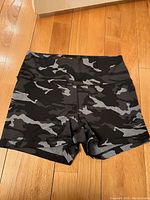 Front view of women's activewear shorts in black and gray camouflage pattern with high waistband lying flat on wooden floor.