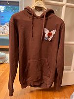 Full front view of brown Hero 5020 hoodie hanging on a hanger in front of a glass door, showing basic style and color.