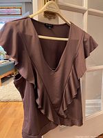 Close-up front view of a brown sleeveless summer top with flutter ruffle details along the front V-neckline, displayed on a hanger in indoor setting.