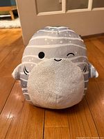 Front view of gray and white striped whale Squishmallow plush toy with winking eye and small fins