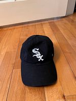 Front view of black Chicago White Sox baseball cap with white embroidered logo, placed on floor.