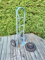 Side view of blue metal dolly with flat base plate, two pneumatic tires installed, exposed rust and wear, extra wheelbarrow tire displayed next to dolly on wooden surface outdoors.
