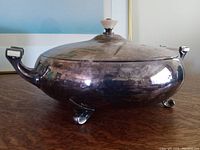 Front and side view of large silverplate lidded tureen with Bakelite handles and knob standing on three rounded feet, showing light tarnish on surface.