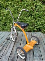 Front and side view of 1970s orange chopper trike with chrome ape hangers and black vinyl seat, showing minor rust and wear.