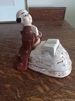 Porcelain figurine of Friar Tuck kneeling next to a textured removable lid shaped as a stone mound with a book on top.
