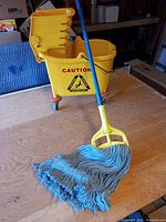 Mop with blue handle and thick blue mop head positioned next to yellow mop bucket with caution sign.