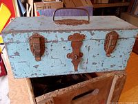 Front view of rectangular metal strongbox showing three rusted metal locking clasps and central escutcheon plate with punched initials 'SM'. Light blue chipping paint throughout.