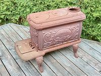Side angle of rusty decorative cast iron garden wood stove on outdoor deck with bushes in background.