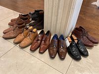 Eight pairs lined up on floor against column: two tan lace-ups, two brown dress shoes, black loafers, maroon chukkas, black oxfords and brown suede boots