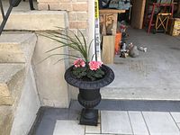 Front view of one black metal urn planter containing soil, pink flowers, and a grassy plant on a paved surface