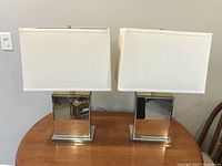 Front view of two modern table lamps with mirrored rectangular bases and white fabric rectangular shades placed side by side on a wooden table.