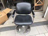 Front view of black padded barbers chair showing seat, backrest, and armrests with chrome metal structure and circular base.