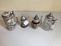 Photo showing four German beer steins of different designs, two ceramic and one pewter, one glass, all with metal lids and handles.