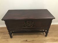 Front view of the cedar chest showing carved wood design and floral fabric underlay on front panel, with turned legs and decorative apron.